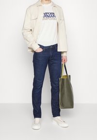 Light beige button-up shirt over a white graphic t-shirt, dark blue jeans, white sneakers, and a green tote bag with a yellow interior.