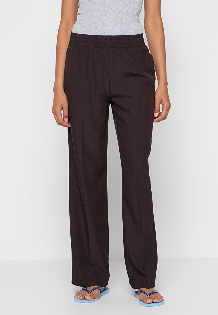 Bruine brede broek met een elastische tailleband, gemaakt van een gladde stof. Gecombineerd met blauwe slippers op een neutrale achtergrond.