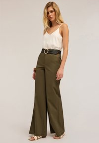 Pantalones de pierna ancha color verde oliva con cintura alta y un cinturón negro tejido que presenta una hebilla dorada circular, combinados con un camisón de seda blanco.