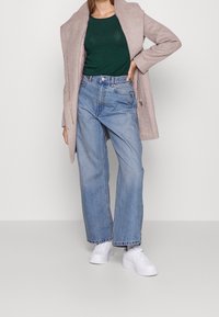 Light beige coat over a green top, paired with high-waisted blue jeans and white sneakers. Casual, relaxed fit with a smooth texture.