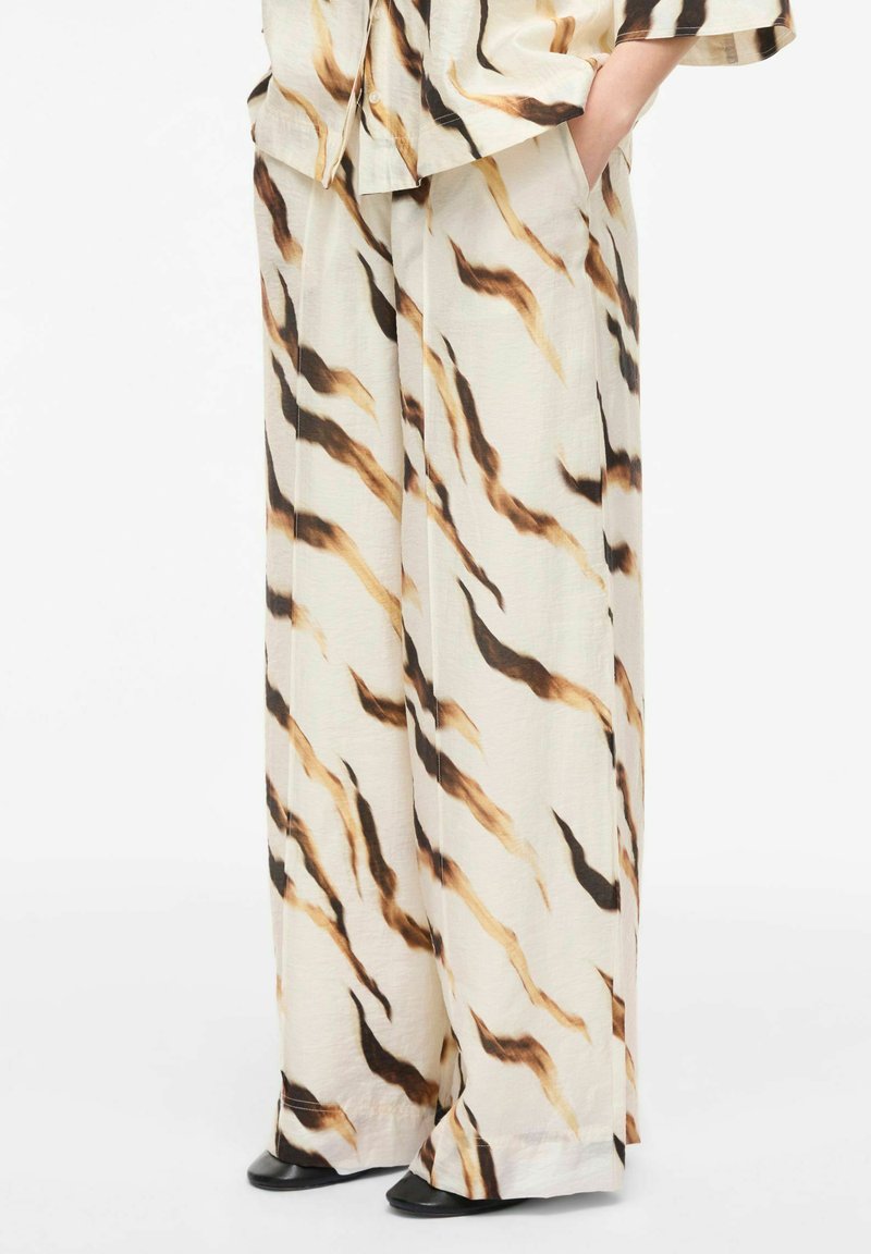 Pantalon ample de couleur crème avec des rayures ondulées abstraites marron et noires, porté avec un haut assorti et des chaussures noires.