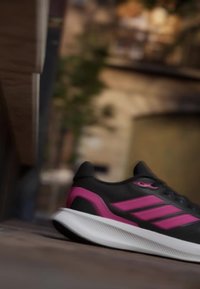 Black and pink athletic shoe with white sole placed on wooden surface, indoor blurred background with plant and window.