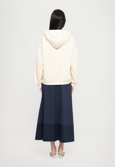 Cream hooded sweatshirt, oversized fit, with ribbed cuffs; paired with a navy maxi skirt featuring a contrasting darker hem.