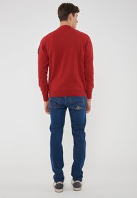 Maglione rosso lavorato a maglia, maniche lunghe, polsini e orlo a costine, abbinato a jeans blu con cuciture decorative sulle tasche posteriori.