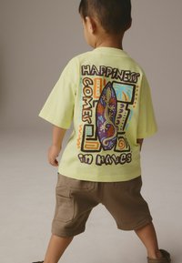 Ljusgrön bomullströja med färggrafisk design som föreställer en surfbräda och texten "Lycka Kommer i Vågorna"; matchad med bruna shorts.