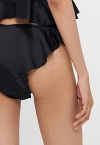 Melnas satīna bikini apakšdaļa ar kraukšķīgiem maliņām un gludu tekstūru. Iespējama redzama ādas tekstūra un stiepšanās zīmes uz gurniem.