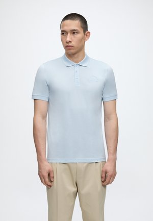 Noor mees, kes kannab helesinist lühikese varrukaga polosärki ja beežeid pükse, seisab valge ühevärvilise tausta ees.