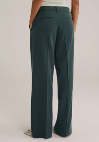 Pantaloni a gamba larga in tessuto verde scuro con una texture liscia. Le caratteristiche includono una vita, tasche laterali e due tasche posteriori.