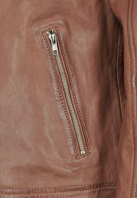 Brun läderjacka med sidofickor med dragkedja och silverdetaljer. Ytan är jämn med synlig ådring och sömdetaljer.