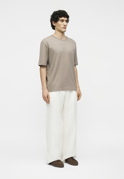 Άνδρας με σγουρά μαλλιά στέκεται φορώντας μια ταμπά κοντομάνικη μπλούζα, φαρδιά λευκά παντελόνια και καφέ παπούτσια μπροστά σε ένα απλό φωτεινό φόντο.