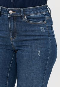 Pogled od blizu na osebo, ki nosi temno modre traper hlače z rahlimi obrabljenimi predeli in temno rebrasto majico, zastrgano v hlače.