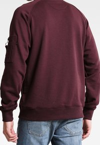 Bordeauxfarbener Pullover aus Baumwollmischung mit geripptem Saum und Bündchen. Verfügt über eine Tasche am linken Ärmel für praktische Zwecke. Sichtbare Nahtdetails.