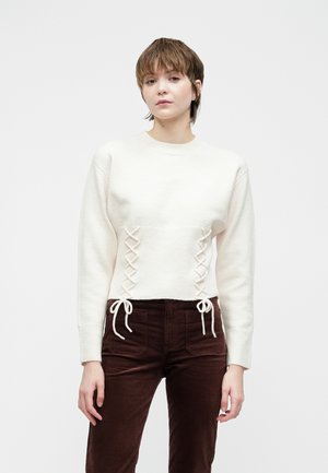 Jeune femme avec les cheveux bruns courts portant un pull en tricot blanc avec un décolleté lacé et un pantalon en velours côtelé marron foncé, se tenant devant un fond uni.