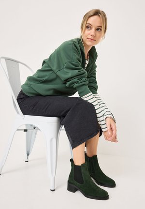 Grüne Wildleder-Stiefeletten mit elastischen Seitenpaneelen und einem leichten Blockabsatz, kombiniert mit einem grünen Sweatshirt und schwarzen weiten Jeans.