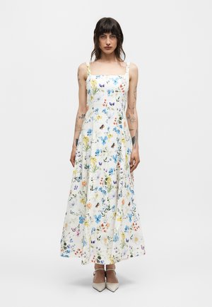 Femme aux cheveux châtains mi-longs portant une robe maxi blanche sans manches avec un imprimé floral et papillons bleu, jaune, rouge, debout immobile.