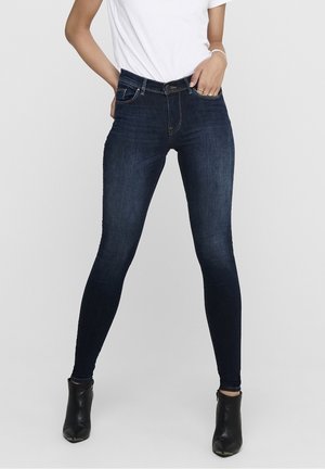 Jeans en denim bleu foncé skinny avec un design classique à cinq poches, des passants de ceinture et un léger délavage. Associés à des bottines noires.