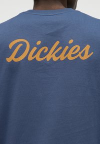 Person trägt ein blaues Hemd mit großem orangefarbenem, kursiv gedrucktem "Dickies"-Schriftzug über dem oberen Rücken.