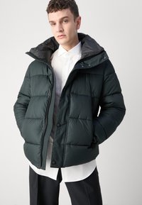 Mørkegrønn pufferjakke med hette, med en quiltet design, frontglidelås og trykknapper, iført over en hvit skjorte.