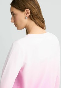Rózsaszín gradient hosszú ujjú póló, amely világos rózsaszínről sötét rózsaszínre vált, kerek nyakkivágással. Ezüst szív alakú fülbevalók díszítik.
