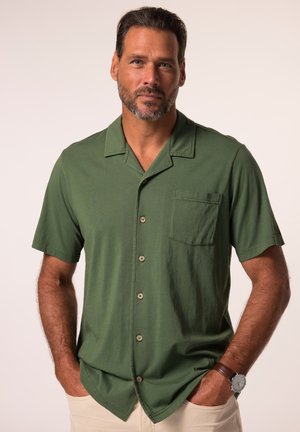 Man met kort donker haar en baard, die een groen kort mouwig overhemd met knopen en beige broek draagt, handen in de zakken.