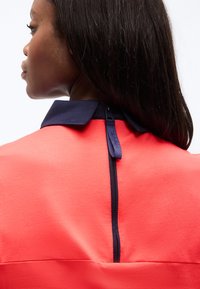 Γυναίκα με σκούρα μαλλιά που φοράει ένα κοραλλί τοπ με ναυτικό γιακά και κατακόρυφο ναυτικό φερμουάρ στο πίσω μέρος.