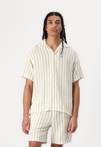 Set di maglietta a maniche corte e pantaloni corti di colore crema leggero con strisce verdi verticali. Design con bottoni frontali e vestibilità rilassata. Tessuto a trama.