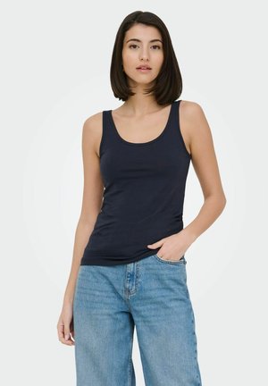 Young woman with straight dark hair wears a navy sleeveless top and light blue jeans, standing with one hand in pocket against a plain background.