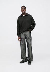 Camisola preta com gola e fecho de correr frontal, combinada com jeans cinzentos. Modelo está em pé, com as mãos relaxadas, usando sapatos pretos.