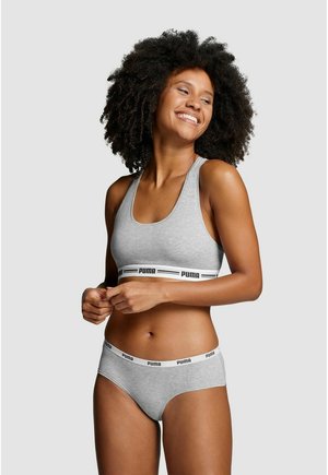 Frau mit lockigem Haar, die lächelt und einen grauen Puma-Sport-BH sowie passende Slips vor hellem, schlichtem Hintergrund trägt.