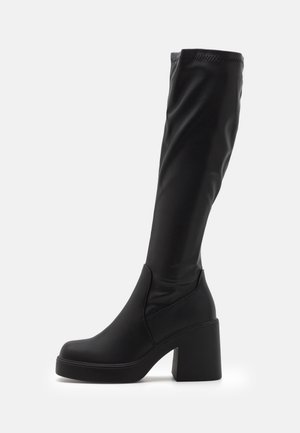 Botte haute en cuir noir avec talon épais et bout rond, s'étendant juste en dessous du genou sur un fond blanc.