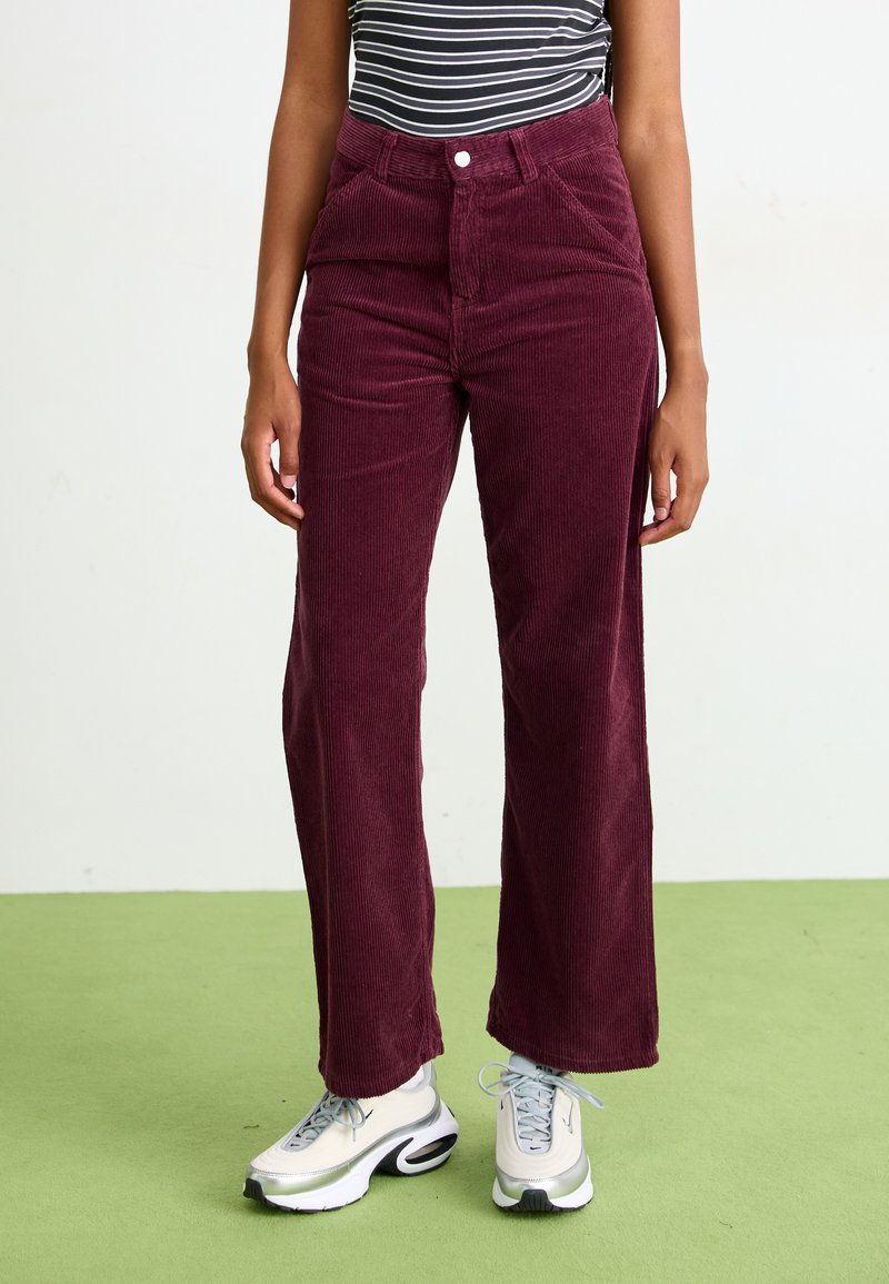 Pantaloni a gamba larga in corduroy bordeaux con superficie testurizzata, vita alta e tasche frontali, abbinati a sneakers sportive bianche e grigie.