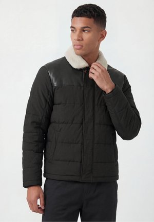 Man wearing black quilted jacket with leather yoke and cream shearling collar, adjusting collar, looking sideways.