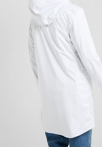 Giacca impermeabile bianca con cappuccio, caratterizzata da una texture liscia e un design semplice, vista da dietro con una vestibilità comoda e maniche lunghe.