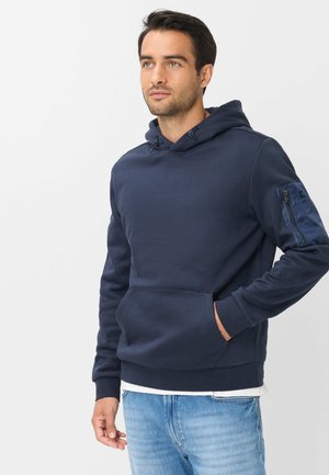 Mann mit kurzen dunklen Haaren, der einen marineblauen Kapuzenpullover mit Reißverschlusstasche am linken Ärmel und hellblaue Jeans trägt.