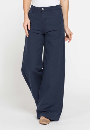Donna che indossa pantaloni blu navy a vita alta e gamba larga abbinati a scarpe piatte intrecciate e un top bianco, in piedi davanti a uno sfondo semplice.