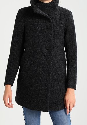 Femme portant un manteau texturé sombre à double boutonnage avec un col montant et des poches latérales, associé à un jean bleu.