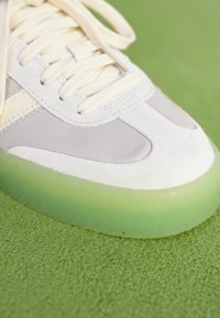Sneaker with a light grey leather upper, white suede accents, cream laces, and a translucent green rubber sole.