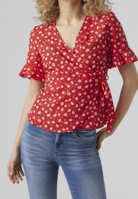Blusa avvolgente rossa a fiori con maniche corte a volantino, caratterizzata da un nodo in vita. Abbinata a jeans attillati blu, creando un look casual.