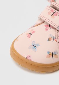 Rozā polsterēts apavi ar krāsainu tauriņu rakstu, Velcro siksnām un brūnas gumijas zoli, kas nodrošina saķeri un komfortu.