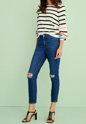 Žena v proužkovaném tričku s dlouhým rukávem, roztrhaných modrých úzkých džínách a černých sandálech na podpatku, stojící na světle zeleném pozadí.