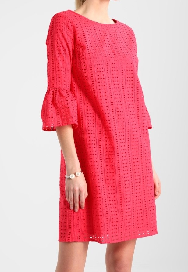 Robe rouge à motif œillets, arrivant au genou, avec des manches trois-quarts évasées, portée par une femme, accessoirisée avec un bracelet en perles.