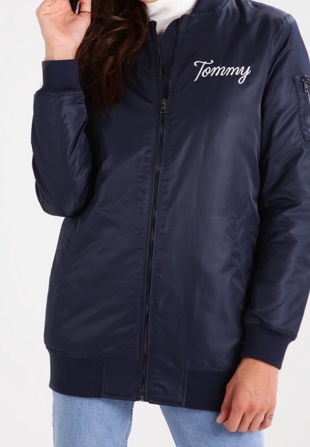 Femme portant une veste bomber bleu marine zippée avec « Tommy » brodé sur la poitrine, associée à un jean bleu clair.