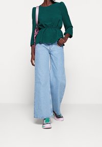 Türkiser Peplum-Oberteil mit langen Ärmeln, kombiniert mit hellblauen weitem Jeans und mehrfarbigen Sneakers. Slingbag in Pink als Akzent.