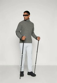 Grå rillede turtleneck sweater, lyse bukser og mørke vandrestøvler. Holder to trekkingstave, bærer solbriller.