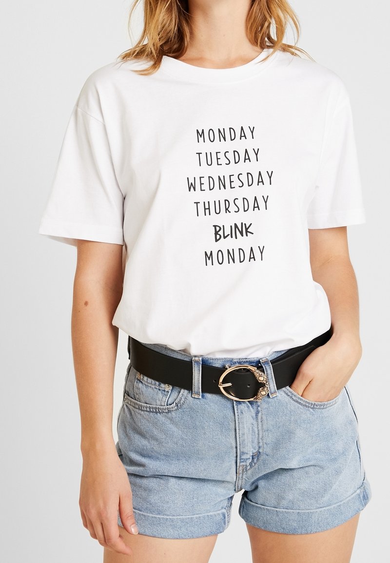 Femme portant une chemise blanche avec le texte listant les jours de la semaine et « BLINK » remplaçant le vendredi, associée à un short en denim clair et une ceinture noire à boucle dorée.