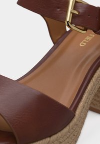 Sandale compensée en cuir marron avec boucle dorée et détail de semelle en corde tressée, gros plan sur la semelle intérieure et la bride.