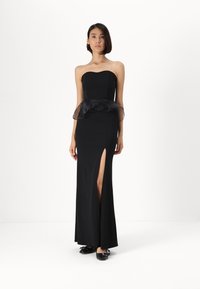 Vestido preto sem alças com corpete justo, volantes pretos transparentes na cintura e uma fenda até a coxa. Modelo usa sapatilhas pretas.