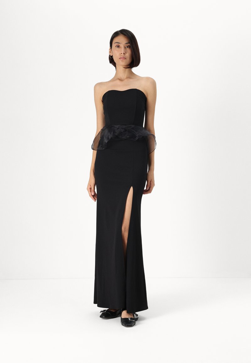 Vestido preto sem alças com corpete justo, volantes pretos transparentes na cintura e uma fenda até a coxa. Modelo usa sapatilhas pretas.