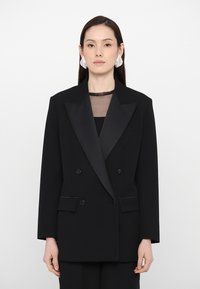Dvojradový čierny blazer so saténovými klopami, jednou náprsní kapsou a dvoma prednými kapsami. Nosený cez priesvitný čierny top.
