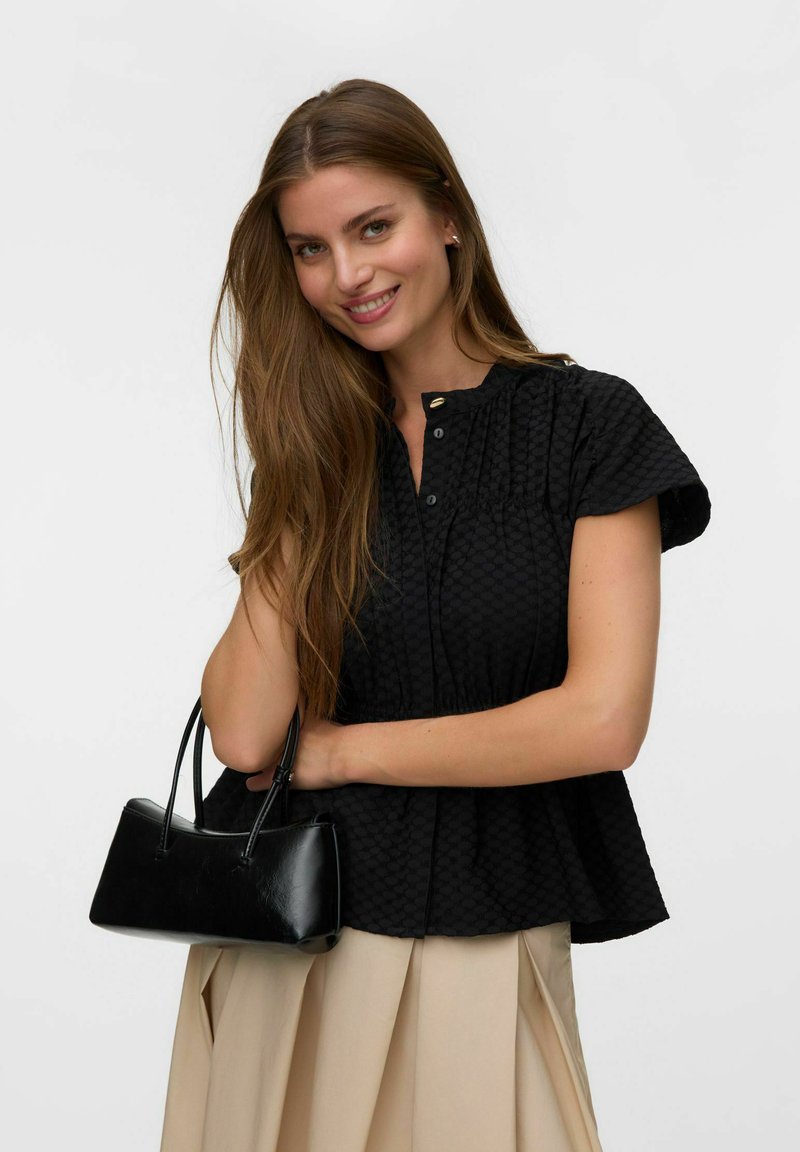 Femme aux longs cheveux bruns portant un chemisier noir texturé et une jupe beige tient un petit sac à main noir et sourit à la caméra.